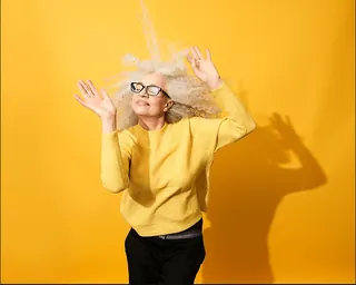 Older woman with white curly hair dancing against a yellow background