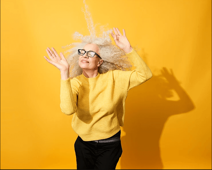 Older woman with white curly hair dancing against a yellow background